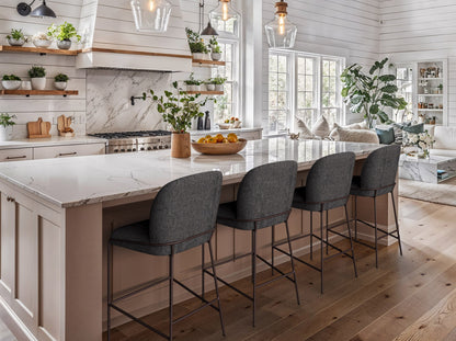 Modern bar stool with a sleek black metal frame, cushioned seat in light gray fabric, and a footrest, set against a minimalist kitchen island with wooden cabinetry and marble countertop.